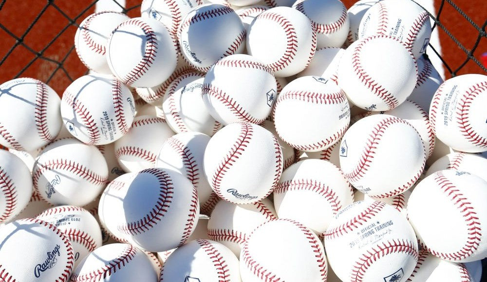 A large pile of white baseballs with red stitching fills a basket, with a net visible in the background.