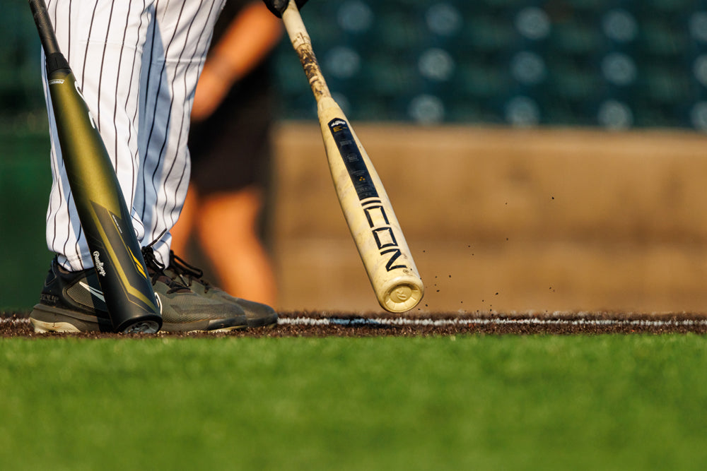 Close-up of a baseball player in black shoes and pinstripe pants holding a 2025 Rawlings Icon (-3) BBCOR Baseball Bat: RB5I3 (DEMO) at the plate, with another black bat nearby on the green grass.