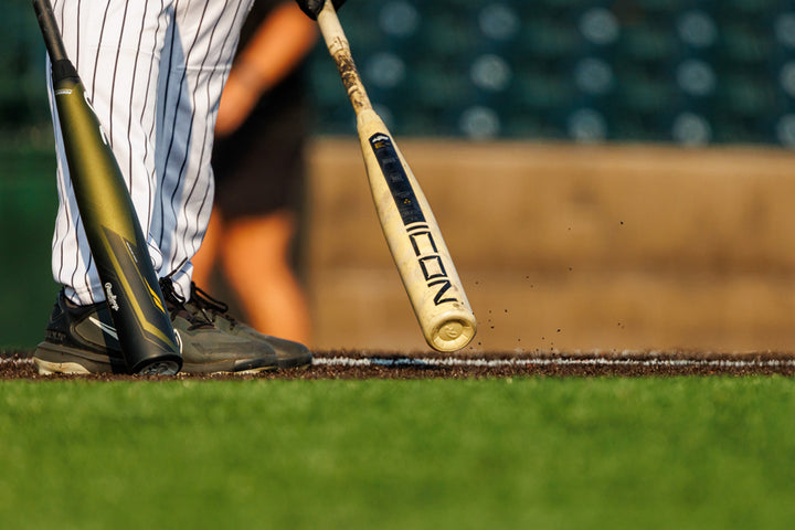 Close-up of a baseball player in black shoes and pinstripe pants holding a 2025 Rawlings Icon (-3) BBCOR Baseball Bat: RB5I3 (DEMO) at the plate, with another black bat nearby on the green grass.
