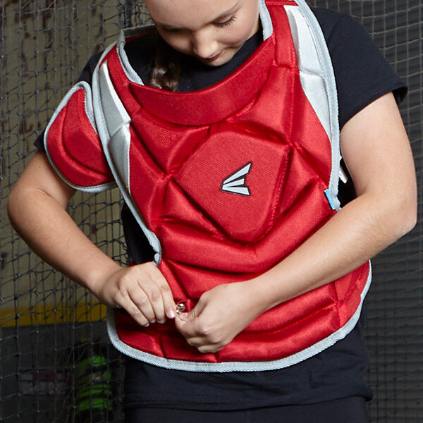 A young person wearing a black shirt fastens a red chest protector, likely for baseball or softball, preparing for practice or a game inside a netted sports facility.