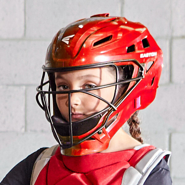 A young softball catcher wearing a red helmet and protective gear looks toward the camera. The background is a gray cinder block wall.