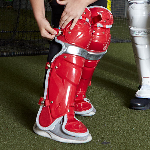 A person is adjusting bright red shin guards with silver accents over black sports pants, standing on artificial turf next to another person wearing silver shin guards.