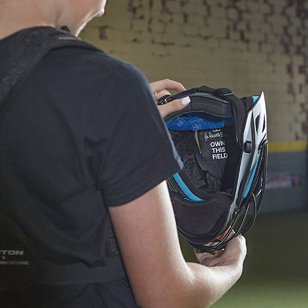 A person holds a black and blue sports helmet, revealing the inside label that reads “OWN THIS FIELD.” The background shows an indoor sports facility with a yellow wall and artificial turf.