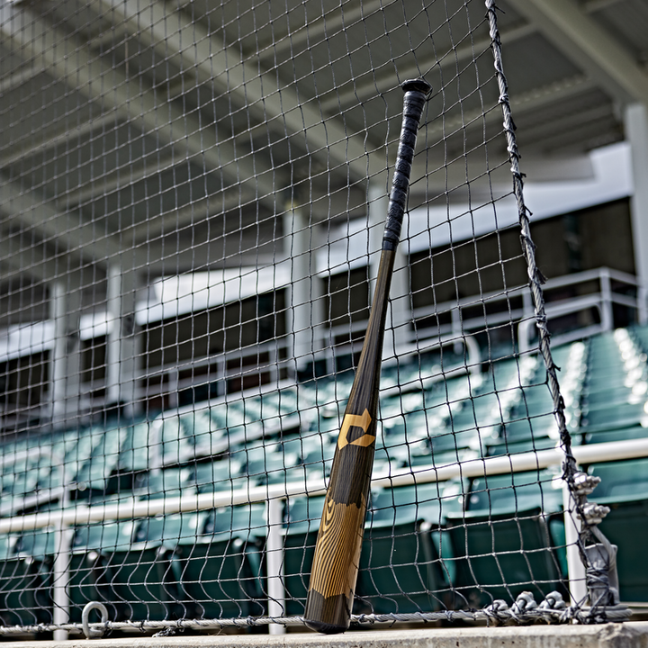 A wooden bat leans on a net before empty green stadium seats, highlighting the contrast between classic wood and the modern DeMarini 2024 Voodoo One (-3) BBCOR Baseball Bat (WBD2461010) seen with a metal railing and stadium roof in the background.