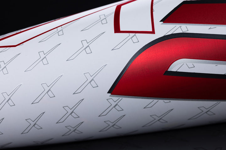 Close-up of a white surface with red and black graphics, bold lettering, and small X patterns—showcasing the one-piece alloy construction and anti-vibration technology of the 2025 Marucci CATX2 (-3) BBCOR Baseball Bat: MCBCX2.