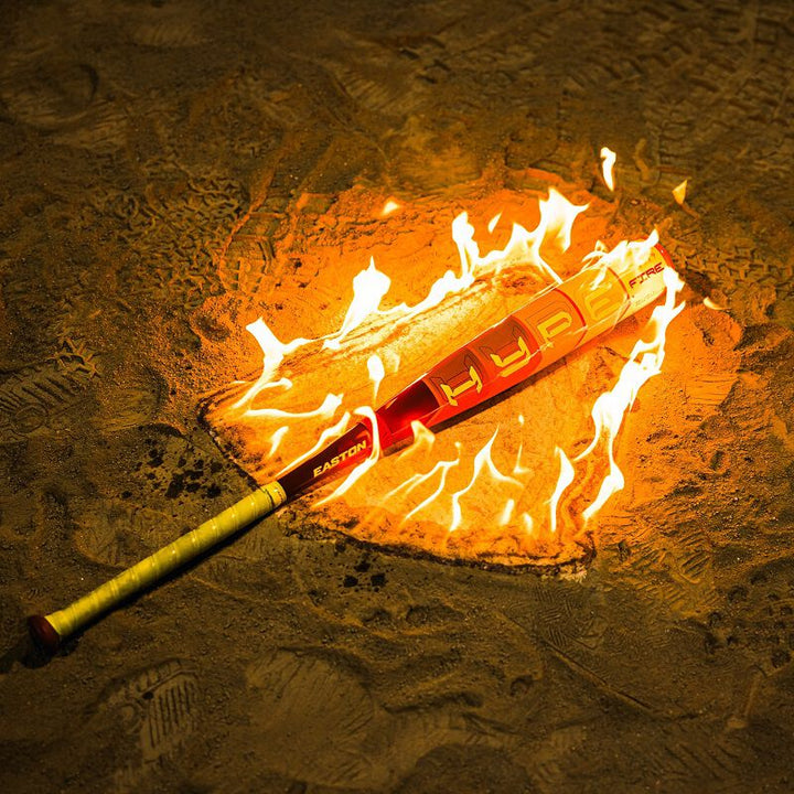 The 2025 Easton Hype Fire JBB (-10) 2 3/4" USSSA Baseball Bat by Easton rests on sandy ground, its label illuminated by orange flames, highlighting the power of Thermo-Composite technology.