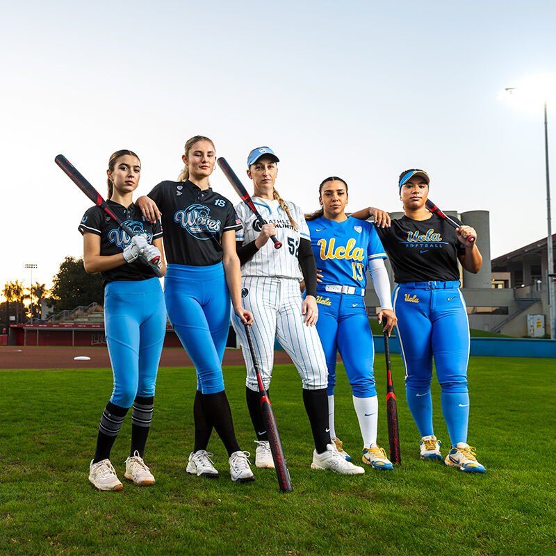 Five female softball players stand on a field holding the 2024 Easton Ghost Advanced (-9) Fastpitch Softball Bat (EFP4GHAD9). They wear UCLA uniforms in blue, black, and white, with stadium lights shining in the background.
