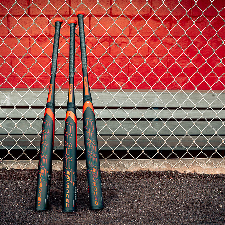 Three black and orange fastpitch bats, including the 2024 Easton Ghost Advanced (-11) by Easton with Double Barrel 3 technology, lean against a chain-link fence in front of a red wall and metal bench on asphalt.