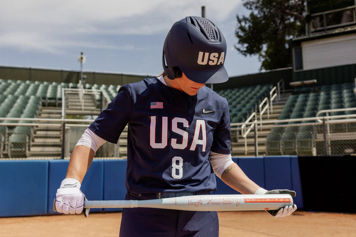 A softball player in a navy blue USA uniform and helmet stands on the field holding an Easton 2025 Ghost Unlimited (-10) Fastpitch Softball Bat (EFP5GHUL10), looking down at it, with mostly empty stands in the background.