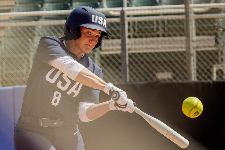 A softball player in a navy USA uniform readies to hit a yellow softball with the 2025 Easton Ghost Unlimited (-9) Fastpitch Softball Bat (EFP5GHUL9), as a chain-link fence and empty seats appear in the background.