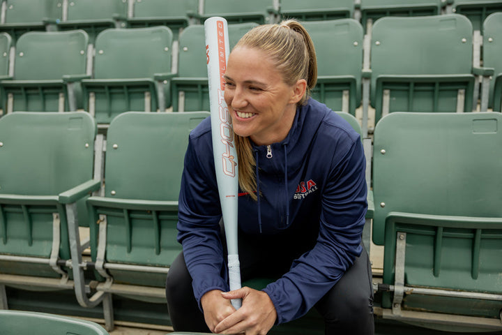 A woman in a navy USA jacket sits smiling in green stadium seats, holding an Easton 2025 Ghost Unlimited (-9) Fastpitch Softball Bat (EFP5GHUL9) vertically in front of her.