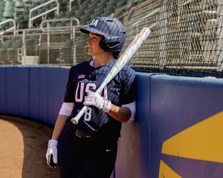 A baseball player in a navy USA uniform and helmet leans on the dugout fence, resting a 2025 Easton Ghost Unlimited (-10) Fastpitch Softball Bat (EFP5GHUL10) by Easton on their shoulder, with empty stadium seats in the background.