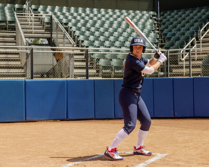 A softball player in a navy USA uniform stands at home plate gripping the 2025 Easton Ghost Unlimited (-11) Fastpitch Softball Bat: EFP5GHUL11 (DEMO) by Easton, with empty stadium seats behind her.