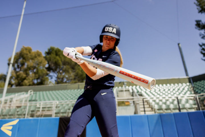 A softball player in a USA helmet and uniform readies to swing the 2025 Easton Ghost Unlimited (-10) Fastpitch Softball Bat (EFP5GHUL10) on a sunny day, with empty green bleachers and trees behind her.