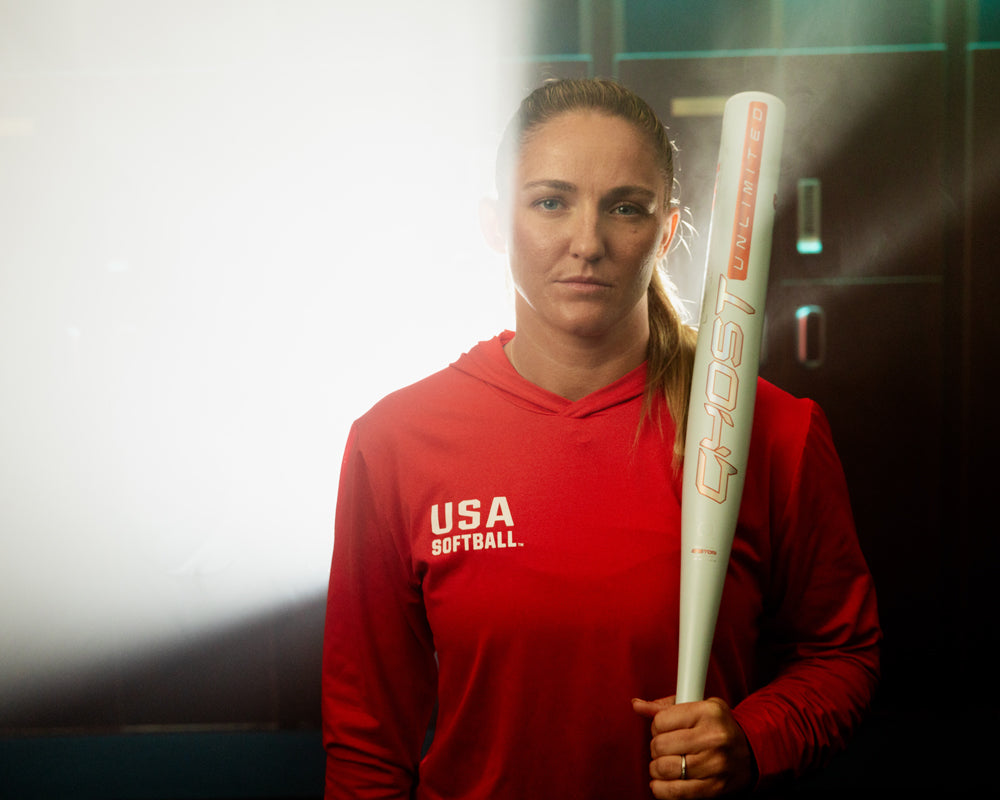 A woman in a red USA Softball hoodie holds the 2025 Easton Ghost Unlimited (-9) Fastpitch Softball Bat (EFP5GHUL9) by Easton, standing in a dimly lit locker room illuminated from the left.