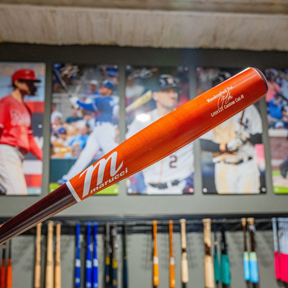 A close-up of the Marucci Francisco Lindor LINDY12 Torpedo Pro Exclusive Maple Wood Bat (MVE5LIND12T) is shown in the foreground, with baseball player posters and rows of colorful Marucci bats displayed on the wall behind it.