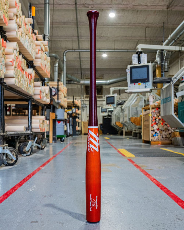 The Marucci Francisco Lindor LINDY12 Torpedo Pro Exclusive Maple Wood Bat (MVE5LIND12T), featuring a red and orange gradient finish, stands upright on the factory floor among machinery, wooden billets, and shelves.