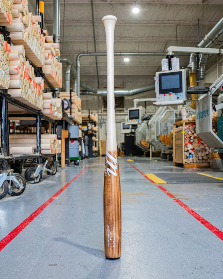 A Marucci AP5 Torpedo Pro Model Maple Wood Bat: MVE5AP5T stands upright on the factory floor, surrounded by machinery and stacks of wood—ideal for power hitters who demand quality from Marucci. Bright lights shine on polished concrete floors.