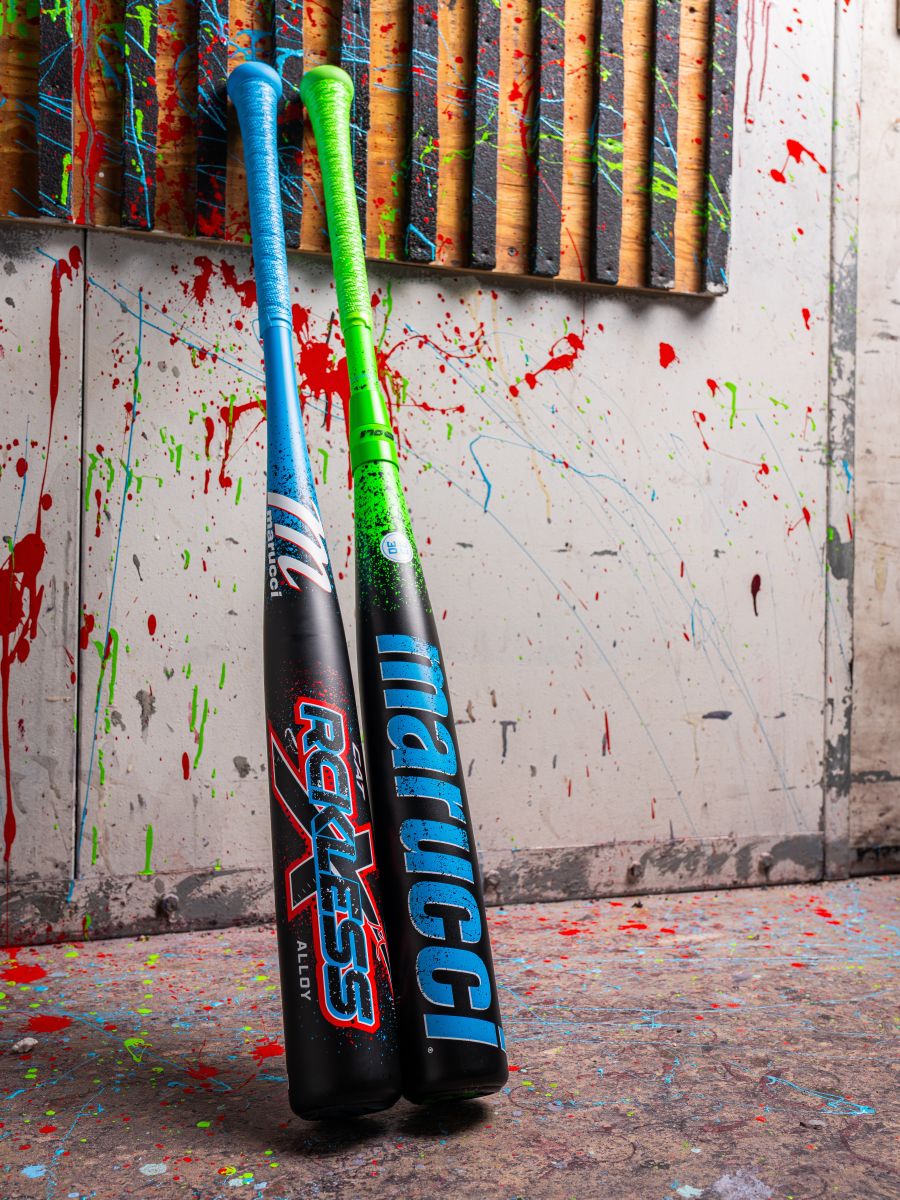 Two vibrant 2026 Marucci CAT X RCKLESS (-3) BBCOR Baseball Bats by Marucci—one blue and one green—lean against a paint-splattered wall with red, green, and blue splashes on the wall and floor.