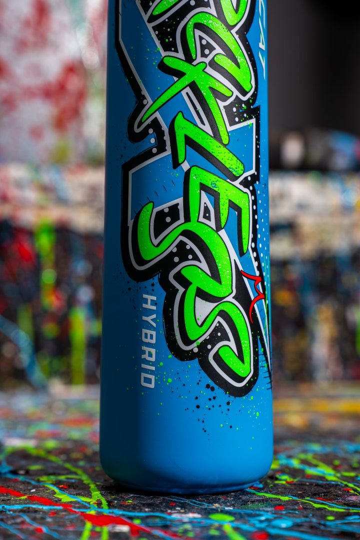A close-up of the blue Marucci CAT X RCKLESS Hybrid (-11) 2 5/8" USA Baseball Bat (MSBCRH11USA) with bold graffiti-style "AXE 52 HYBRID" lettering, set against a vibrant abstract painted backdrop.