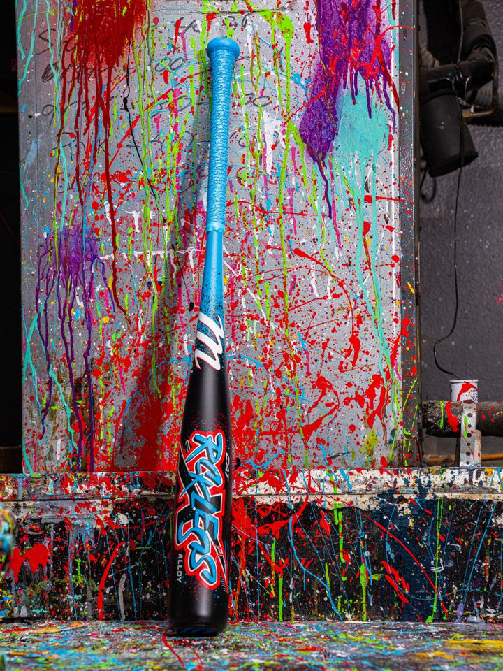 A Marucci CAT X RCKLESS (-8) USSSA Baseball Bat stands upright against a wall splattered with red, blue, purple, and green paint, creating a vibrant abstract backdrop.