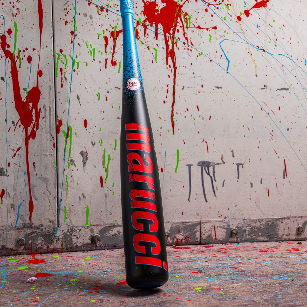 A Marucci baseball bat with a blue handle and black barrel featuring red lettering stands upright against a wall splattered with colorful paint. The floor is also covered with paint splatters.