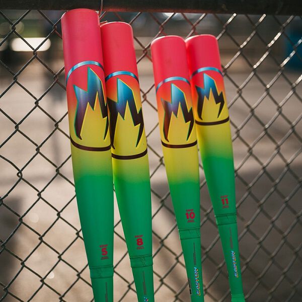 Four colorful USA certified bats with red, yellow, and green gradients plus flame graphics rest against a fence in bright sunlight. The standout is the 2026 Easton Hype Fire (-11) 2 5/8" USA Baseball Bat by Easton, with numbered handles.