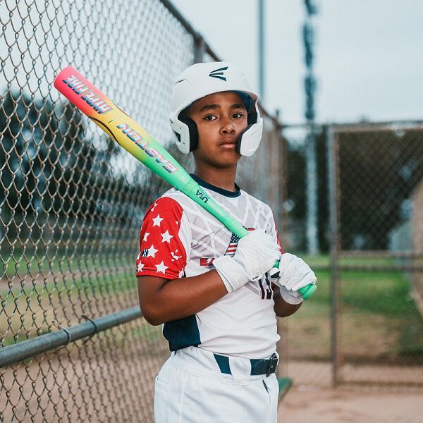 A young baseball player in a white uniform and helmet stands on a field holding an Easton 2026 Hype Fire (-11) 2 5/8" USA Baseball Bat (EUS6HYP11), looking confidently at the camera, with a chain-link fence in the background.
