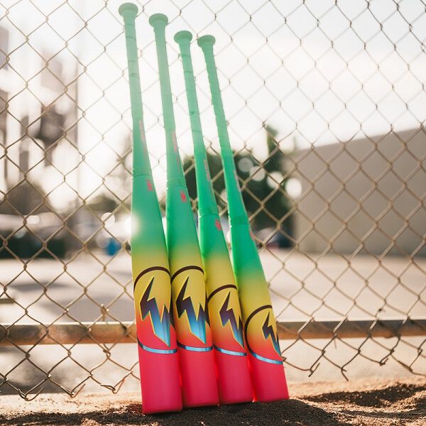 Four brightly colored baseball bats, including the 2026 Easton Hype Fire (-11) USA Baseball Bat by Easton with a green to yellow gradient and flame designs, stand upright against a chain-link fence on a sunny day.