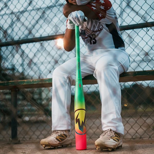 A baseball player in a white uniform sits on a bench behind a chain-link fence, resting his arms on an Easton 2026 Hype Fire (-11) USA Baseball Bat (EUS6HYP11) with vibrant green and red lightning bolt graphics. Only his lower face and body are visible.