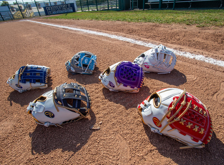 Six baseball gloves, including a Rawlings Liberty Advanced Color Sync 12" Fastpitch Softball Glove (RLA120-3WNG), are laid out on a dirt infield near the white foul line, with a fence and Howell sign in the background.