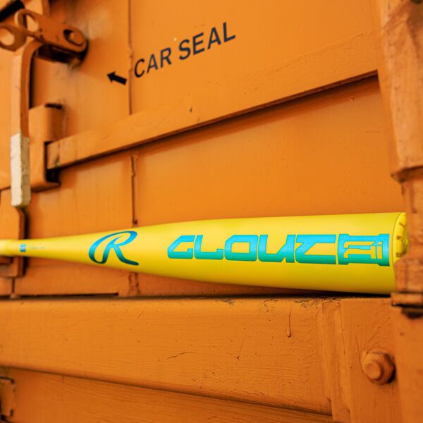 A yellow and blue Rawlings Clout AI (-3) BBCOR Baseball Bat (RBB6C3) rests horizontally on an orange metal surface by a door marked "CAR SEAL," with a black arrow pointing left.