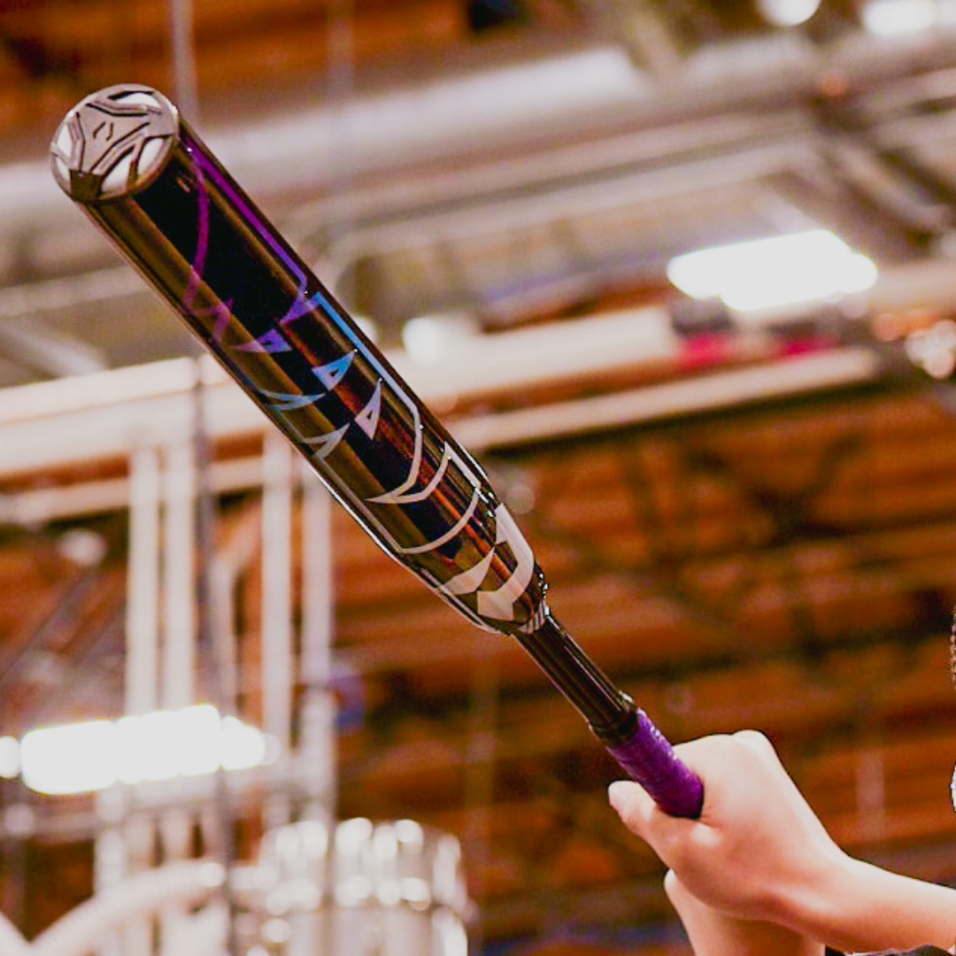 A person holds a DeMarini 2026 Prism+ (-11) Fastpitch Softball Bat (WBD2636010), black and purple, poised to swing, with an indoor industrial background softly blurred.