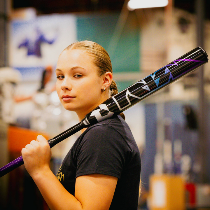 A young woman with blonde hair pulled back holds the 2026 DeMarini Prism+ (-11) FE Fastpitch Softball Bat (WBD2636010) over her shoulder, gazing confidently at the camera in an indoor setting. She wears a dark t-shirt.