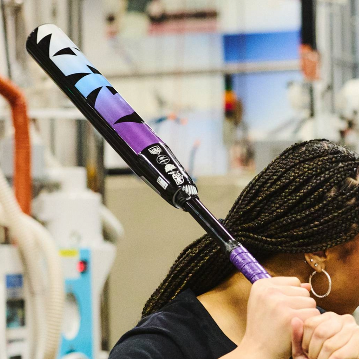 A person with braided hair holds a DeMarini 2026 Prism+ (-11) FE Fastpitch Softball Bat (WBD2636010 DEMO), featuring a Continuous Fiber Composite Barrel, over their shoulder in an indoor workshop or gym. Their face is not visible.