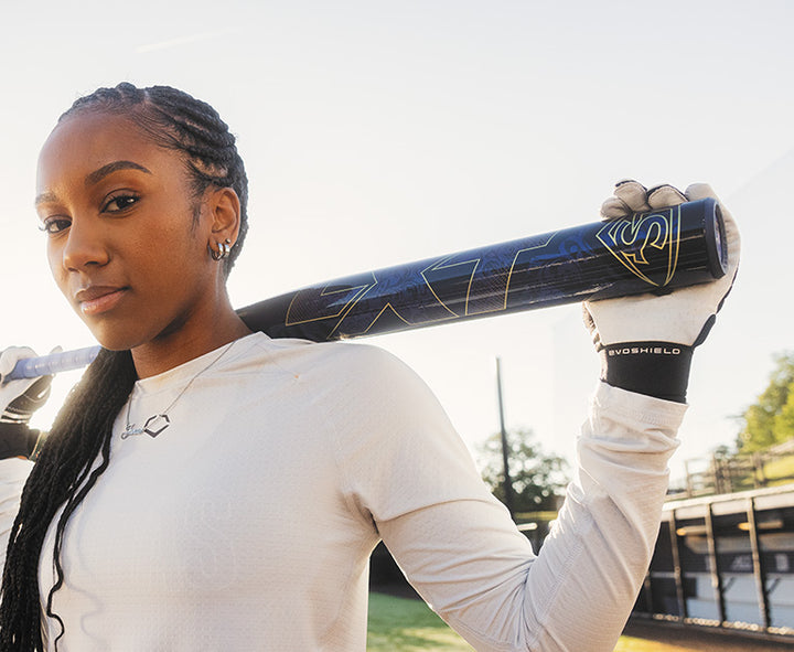 A young woman with braided hair in a white shirt and batting gloves holds the 2025 Louisville Slugger LXT (-9) Fastpitch Softball Bat (WBL2995010) over her shoulders, standing on a sunlit softball field.