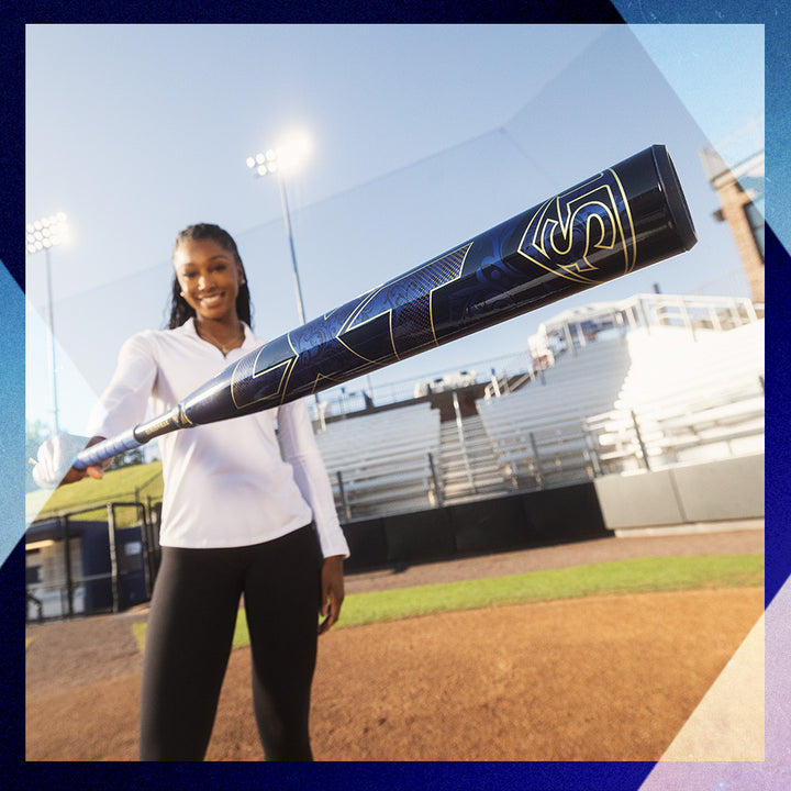 A smiling woman in a white shirt and black pants stands on a baseball field, holding the Louisville Slugger 2025 LXT (-11) Fastpitch Softball Bat (WBL2993010) toward the camera, with empty bleachers and stadium lights behind her.
