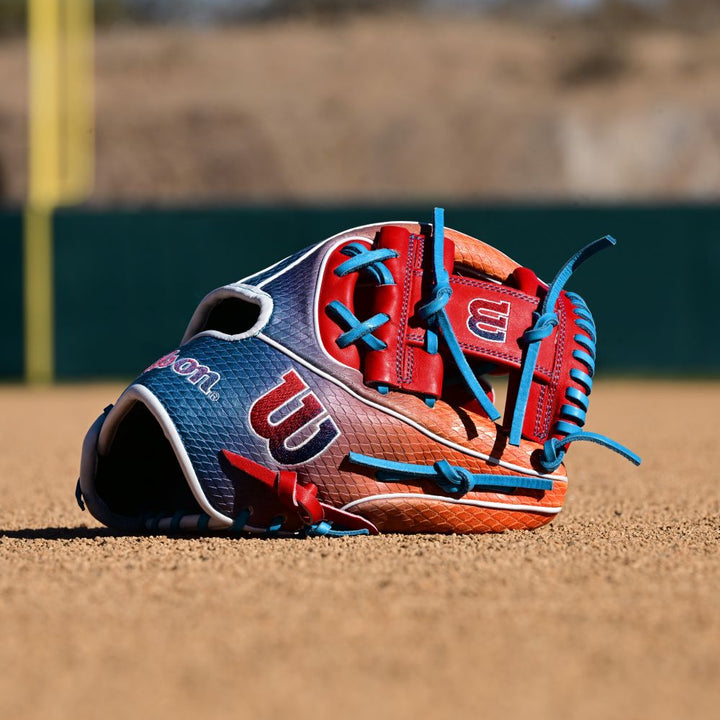 A Wilson A2K 1786SS 11.5" Baseball Glove (WBW103290115) in blue, red, and white with blue laces and SuperSnakeSkin material rests on the dirt; blurred green fencing and a yellow foul pole are visible in the background.