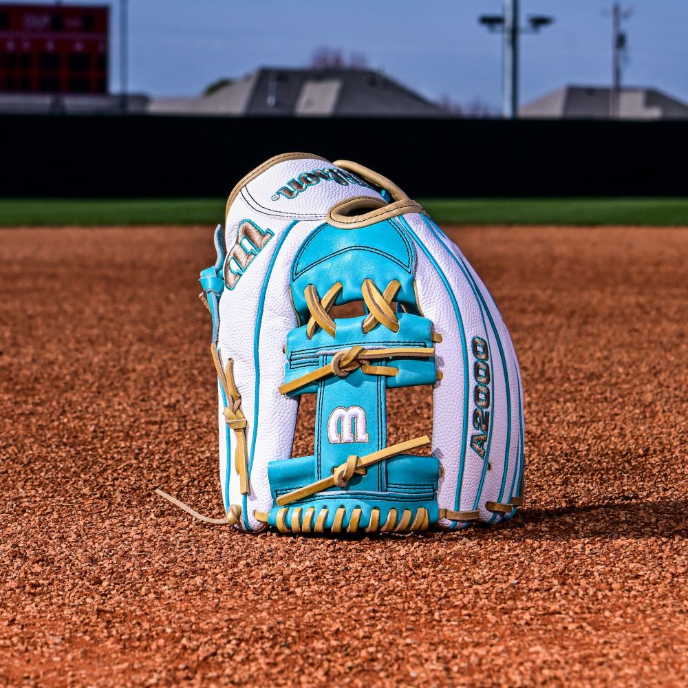 The Wilson A2000 H12SS 12" Fastpitch Glove (WBW10329812), in white and teal with tan laces, stands upright on the infield dirt, Pro Stock leather catching the light, with an outfield fence, scoreboard, and distant buildings blurred behind it.