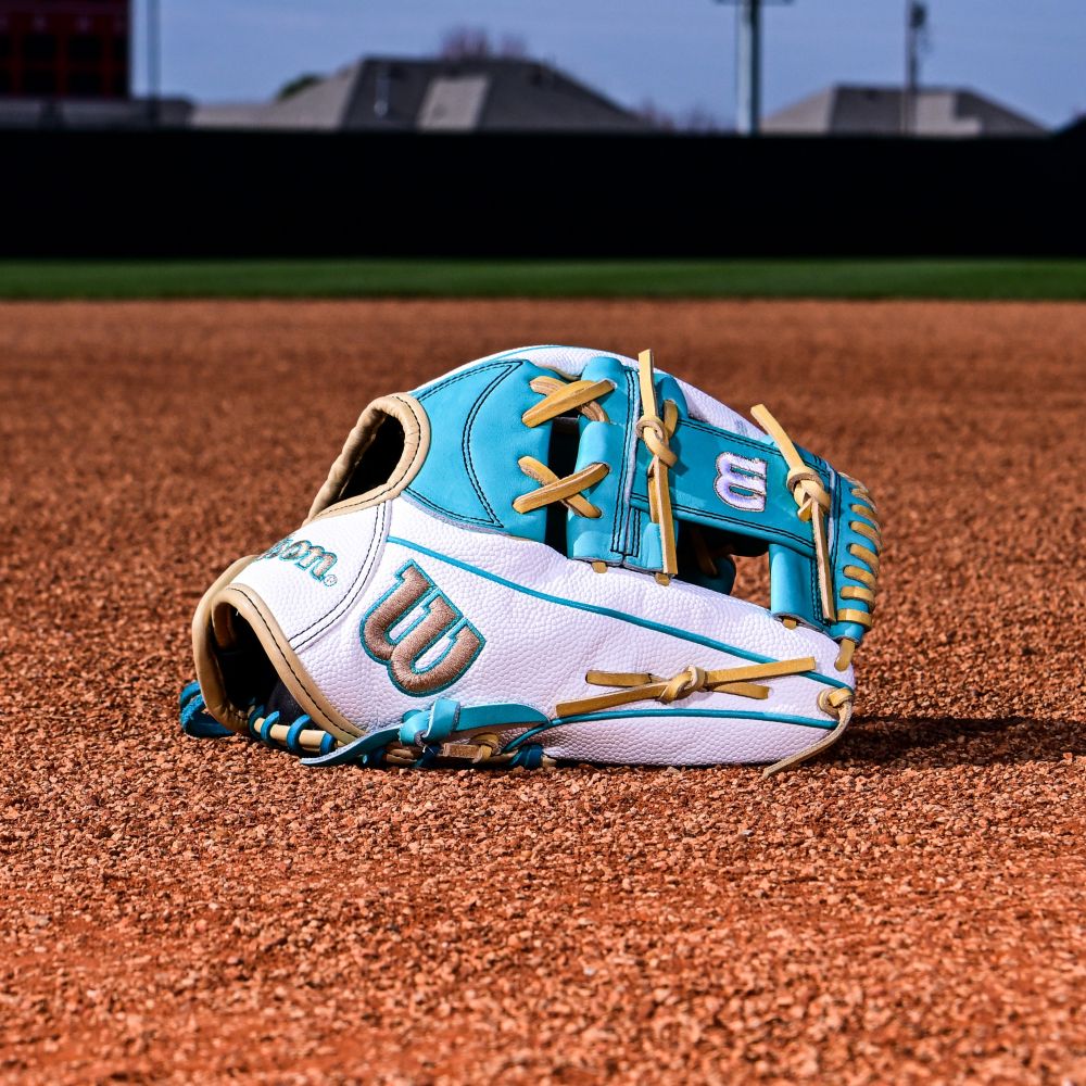 A Wilson A2000 H12SS 12" Fastpitch Glove (WBW10329812) by Wilson rests on the brown infield dirt, with a grassy outfield and blurred buildings visible in the background.