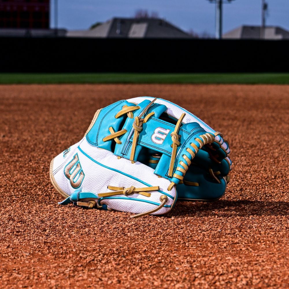 The Wilson A2000 H12SS 12" Fastpitch Glove (WBW10329812) rests on infield dirt, its teal and white Pro Stock leather and tan laces standing out against the grass and distant buildings.
