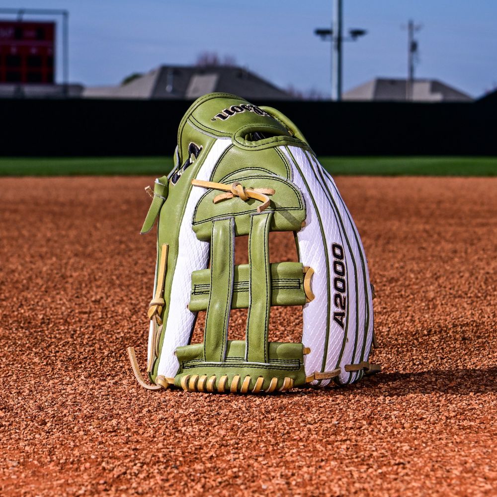 A Wilson A2000 FP50SS 12.25" Fastpitch Glove (WBW1033001225) stands upright on the infield dirt of a baseball field, with green grass and stadium structures in the background.