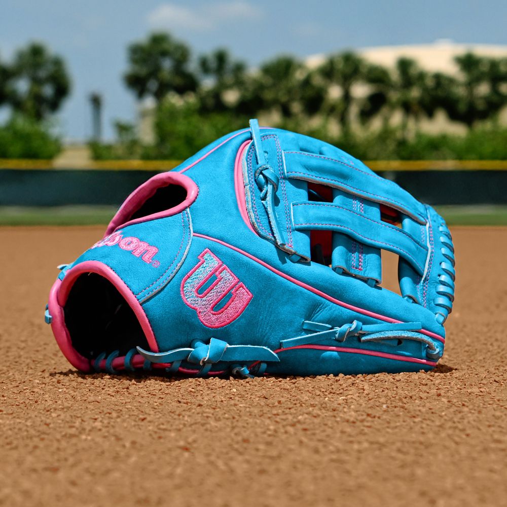 A Wilson A2000 EDLC44 12" Elly De La Cruz Gameday Baseball Glove (WBW10345212) lies on the brown infield dirt of a baseball field, with green grass and trees blurred in the background.