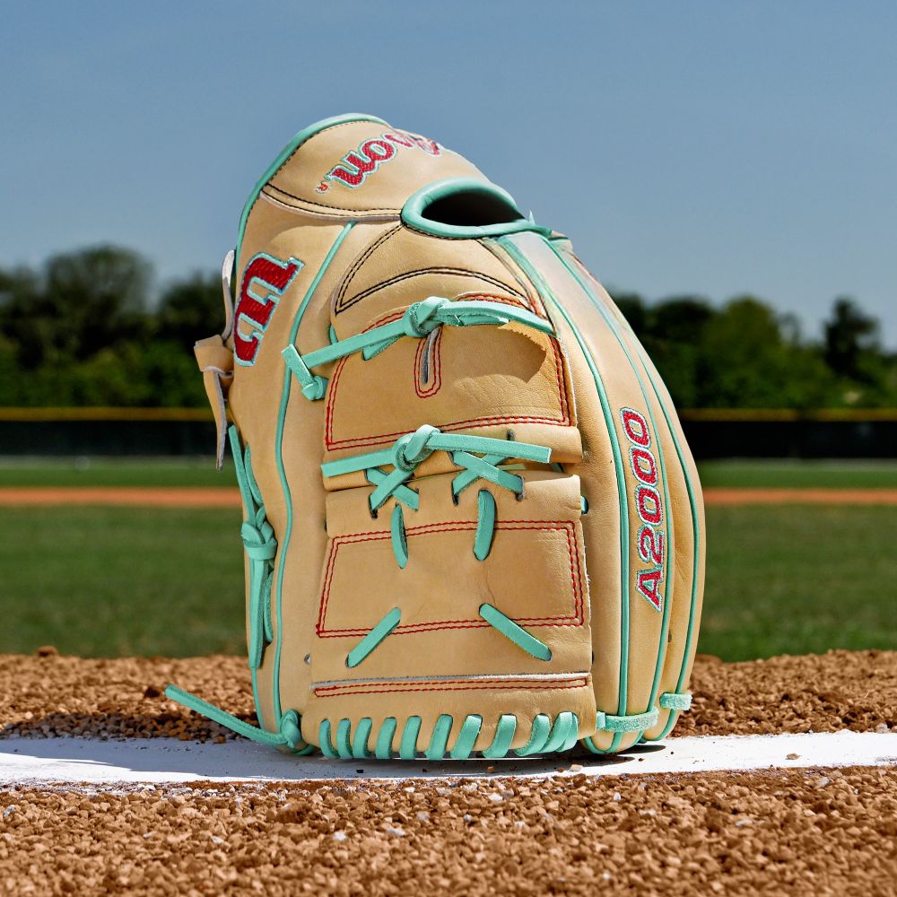 A Wilson A2000 B12 12" Baseball Glove (WBW10349812) stands upright on a pitcher’s mound with green grass, trees, and a baseball field in the background under a clear blue sky.