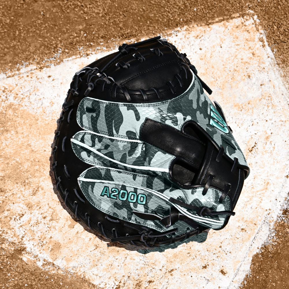 A Wilson A2000 AW28 34" Austin Wells Gameday Catcher's Mitt (FALL 2025, WBW10349434) rests on home plate, its camouflage pattern blending with the dirt.