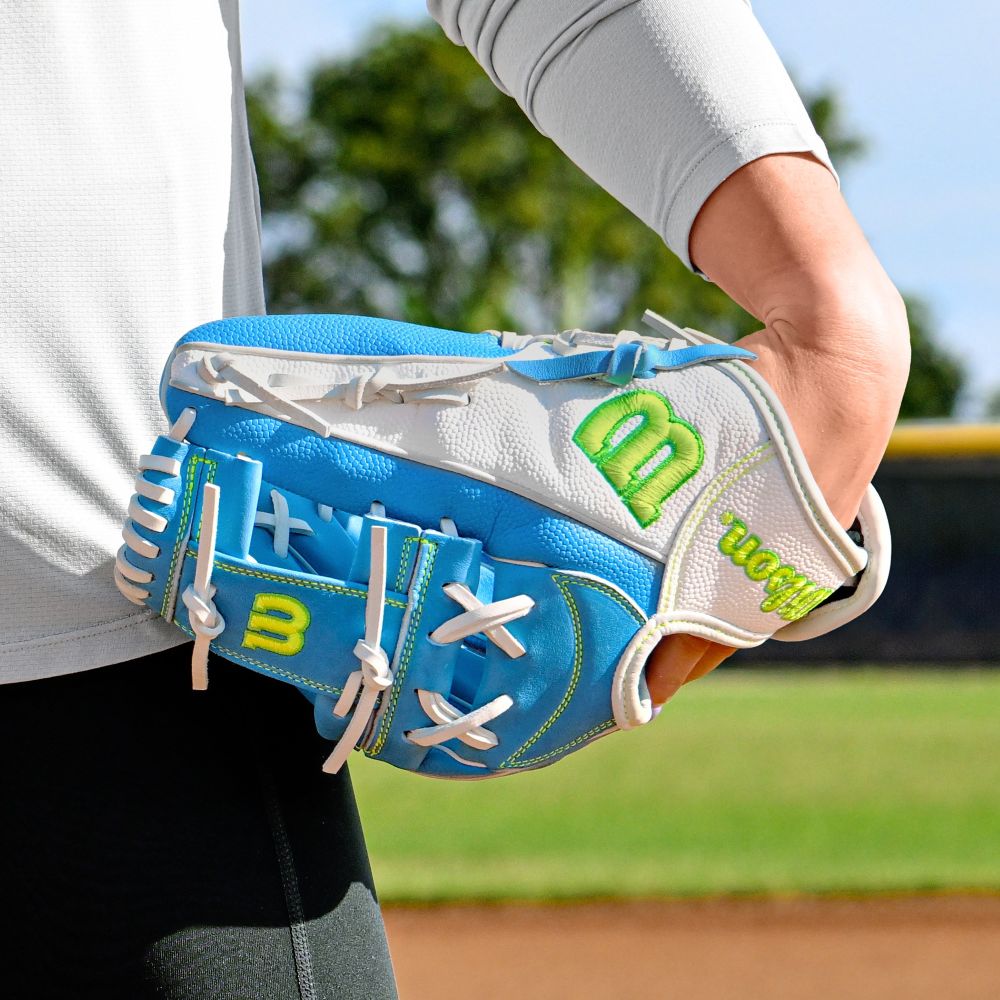 A close-up of a person holding the Wilson A2000 H12SS 12" Fastpitch Glove (WBW10348612), blue and white with green and yellow accents, on a baseball field with trees and grass in the background.