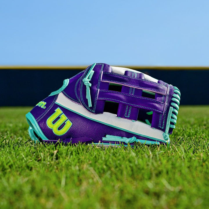The Wilson A2000 FP50SS 12.25" Fastpitch Glove (FALL 2025) features a purple, teal, and white Pro Stock leather design with a yellow W logo, shown on green grass under a blue sky with a blurred fence in the background.