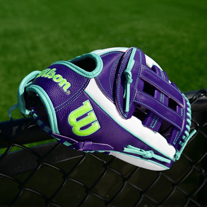 A Wilson A2000 FP50SS 12.25" Fastpitch Glove in purple, teal, and white with neon green accents and Pro Stock leather rests on a black chain-link fence with green grass behind it. Brand: Wilson. Model: WBW1034891225 (Fall 2025).