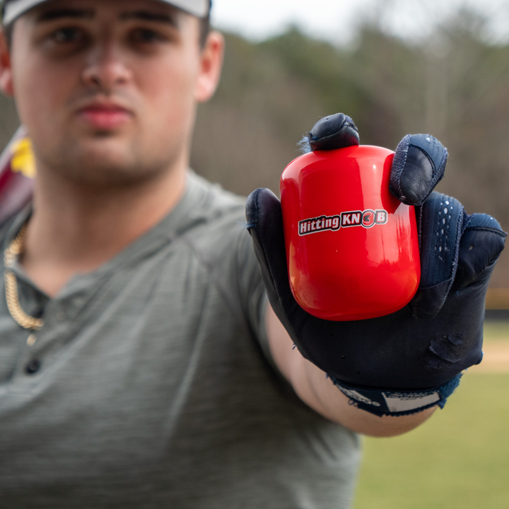 A baseball player wearing a glove holds out The Hitting Knob Bat Weight by Hitting Knob toward the camera, a red baseball training aid designed to improve bat speed, with a blurred outdoor background.