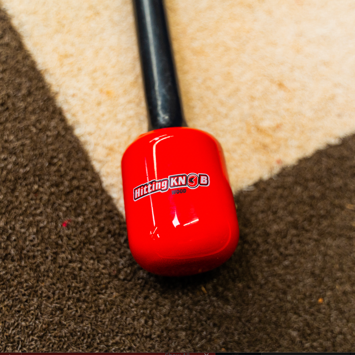 The Hitting Knob Bat Weight by Hitting Knob, featuring a black handle and bright red square end, rests on a brown and beige patterned carpet. This baseball aid is designed to help improve bat speed during practice.